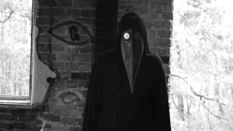 11B X 1371 — Black-and-white photograph of a figure in plague doctor costume with beaked mask and dark robes before a graffitied brick wall.