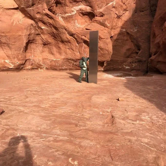 2020 Metal Monoliths — Metal monolith alongside a camouflaged figure in a red rock canyon, showing scale against the structure.