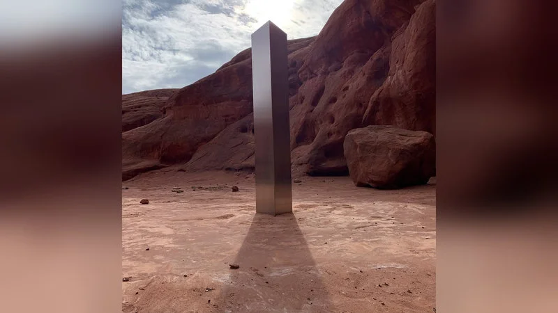2020 Metal Monoliths — Metal monolith standing prominently in red rock canyon with dramatic natural formations, faded edge composition.