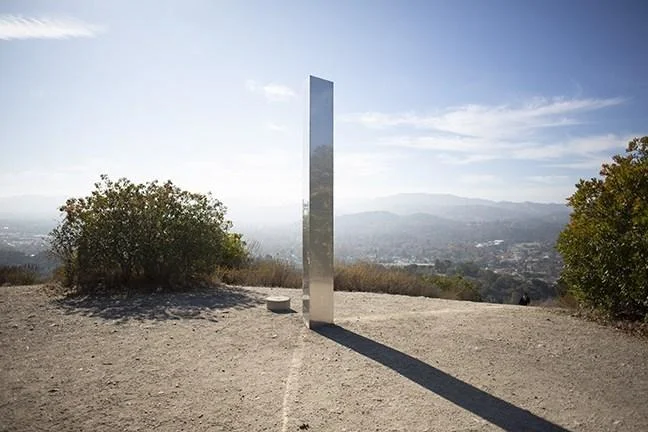 2020 Metal Monoliths — Polished metal monolith standing upright on a hillside overlooking an urban landscape with mountains.