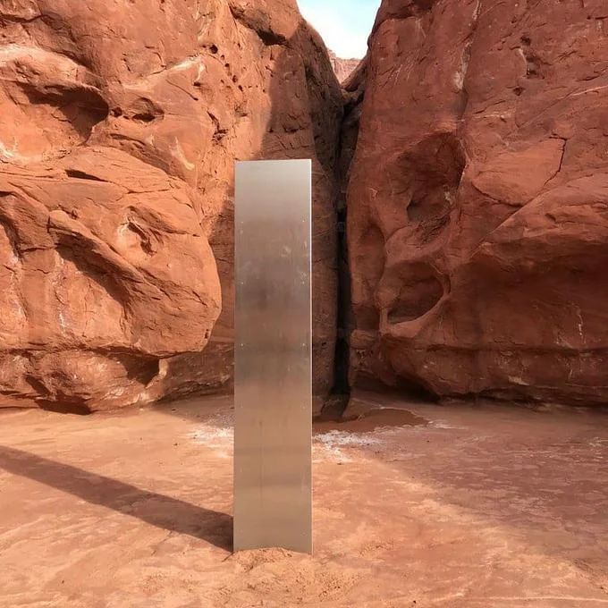 2020 Metal Monoliths — Tall reflective metal monolith standing vertically between towering red canyon rock formations in the desert.