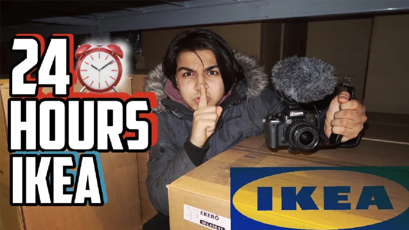 24 Hour Fort Challenge — Person making 'shush' gesture while hiding among cardboard IKEA boxes with camera equipment at their side.