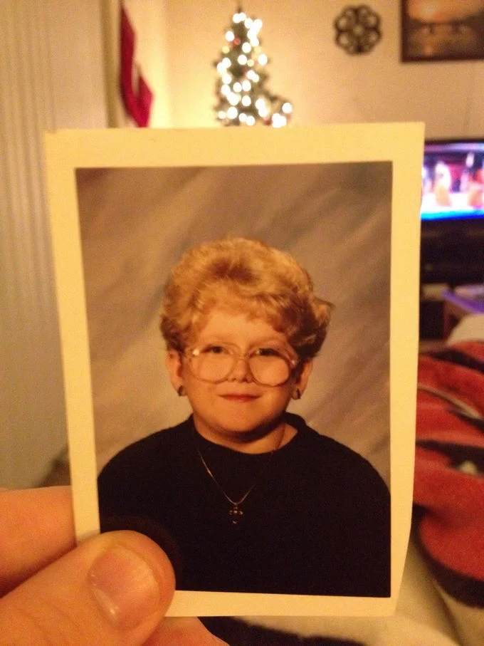 60 Year Old Girl — A portrait photograph of a young girl with short reddish hair, large eyeglasses, and a black shirt against a neutral beige background, the
