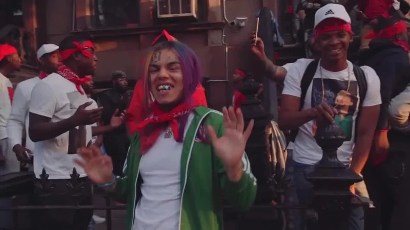 6Ix9Ines Gummo — A still from 6ix9ine's 'Gummo' music video featuring the rapper with vibrant pink and purple hair, wearing a red hood and green jacket in an