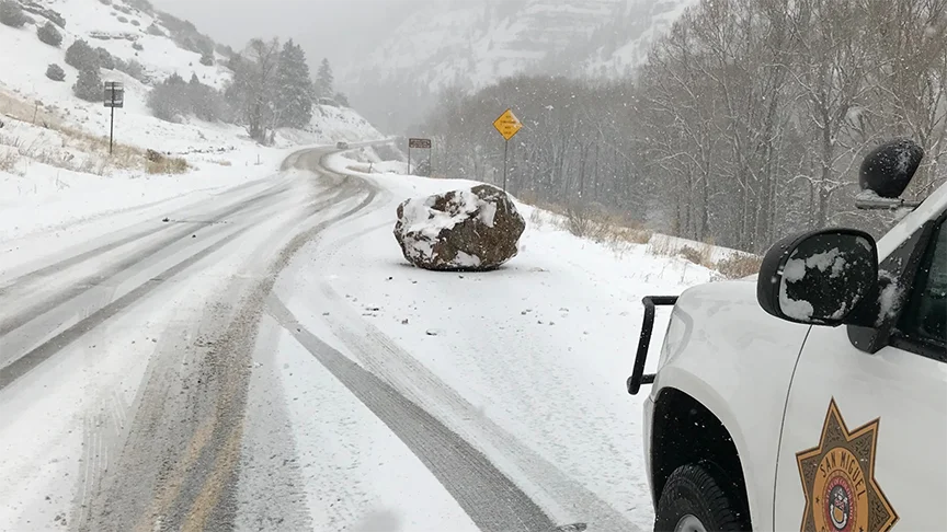 A Large Boulder The Size Of A Small Boulder — Snow-covered boulder blocking a snowy highway next to a San Miguel Sheriff SUV