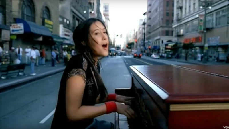 A Thousand Miles Making My Way Downtown — Vanessa Carlton playing an upright piano on a bustling downtown street lined with urban buildings, looking back over