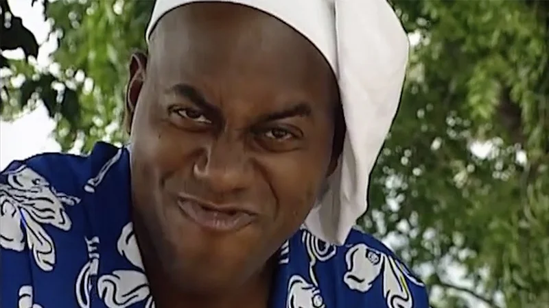 Ainsley Harriott smirking in a blue floral shirt and white headscarf outdoors