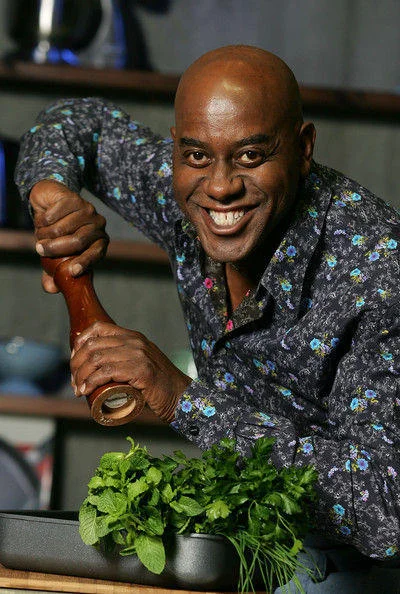 Ainsley Harriott grinning while holding a wooden pepper grinder over herbs