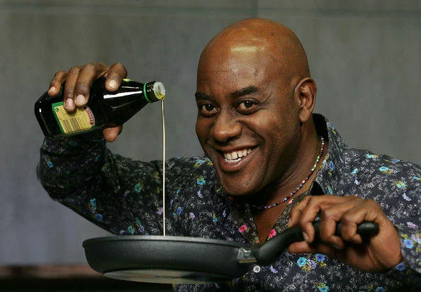 Ainsley Harriott smiling while pouring olive oil into a frying pan