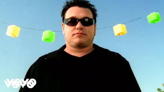 All Star / Shrek — Smash Mouth singer Steve Harwell in the All Star music video, wearing sunglasses under colorful cube string lights