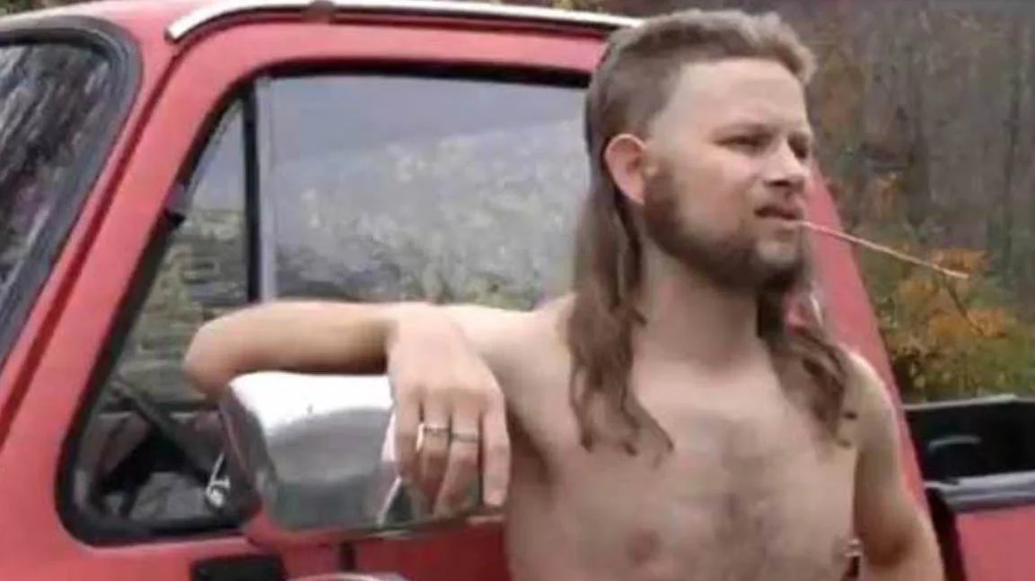 Almost Politically Correct Redneck — Shirtless man with long mullet hair and beard leaning on a red pickup truck with straw in his mouth