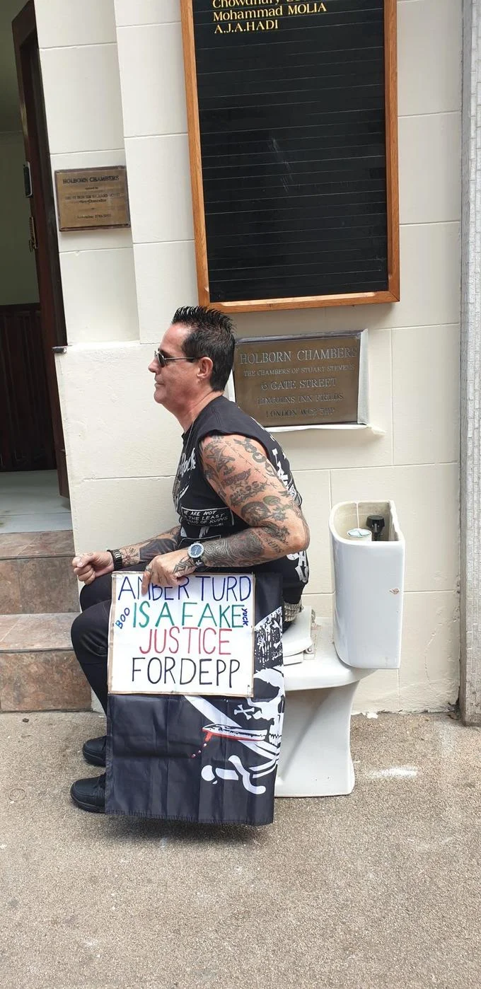 Amber Heard Revenge Poop — A tattooed man seated on a toilet outside a courthouse holding a hand-made sign reading 'Amber Turd is a Fake, Justice for Depp'.