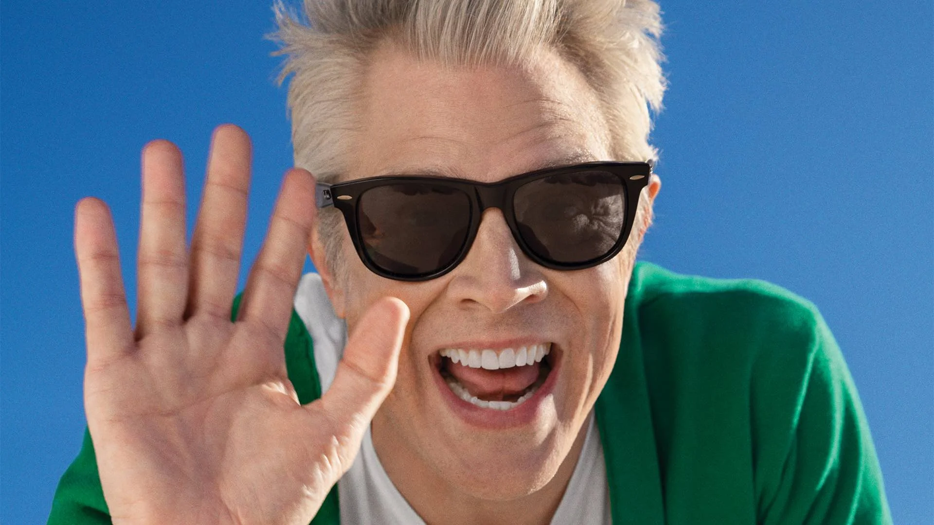 And Welcome to Jackass — Johnny Knoxville in sunglasses and green jacket smiling and waving against a blue sky