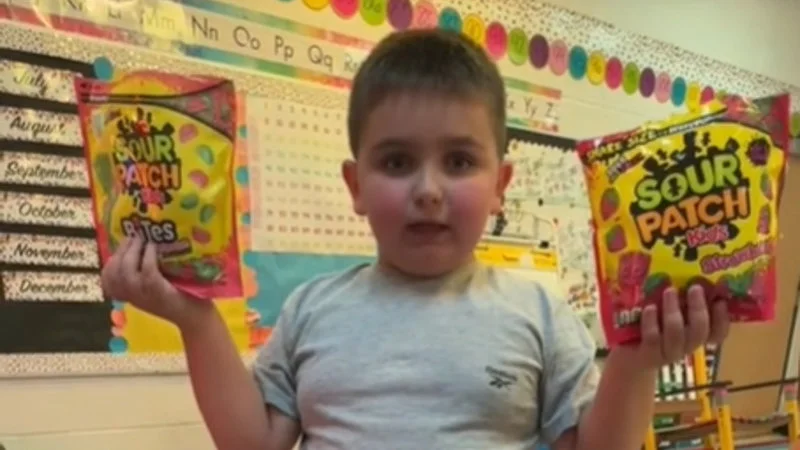 Anderdingus — A young child in a classroom setting holds up two bags of Sour Patch Kids candy with colorful decorations in the background.