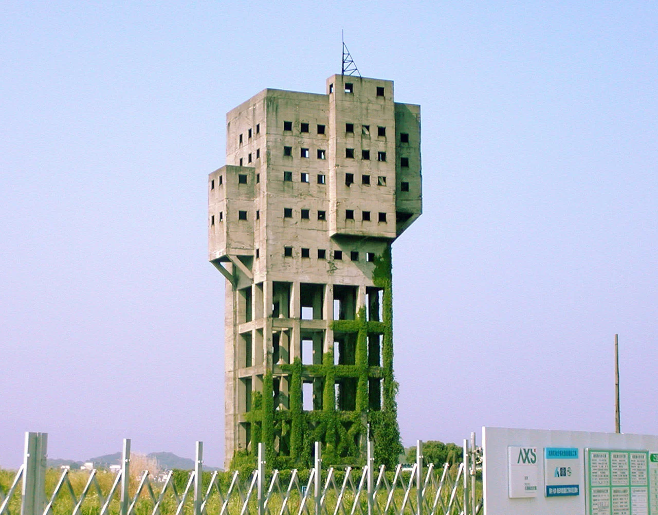 Anti-Zombie Fortress — Tall concrete tower building with grid of small square windows, overgrown with ivy at its base in a field.
