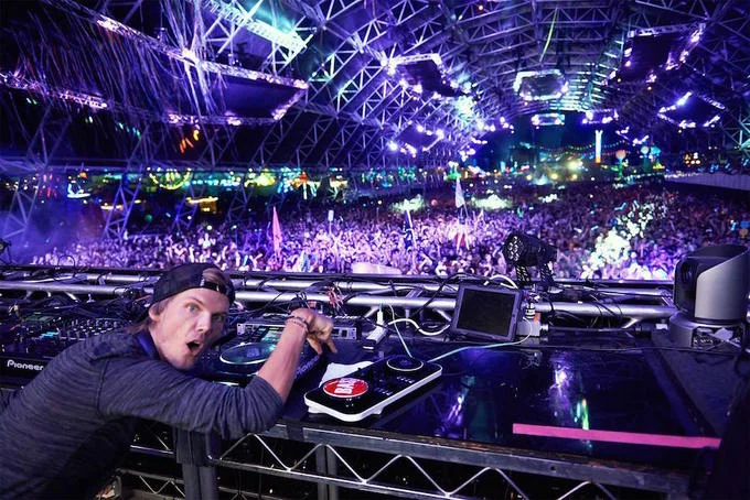 Avicii DJing at a large festival with purple lights and crowd