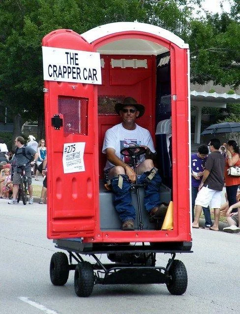 Awful Taste, But Great Execution — Man driving a motorized vehicle built to look like a red portable toilet in a parade