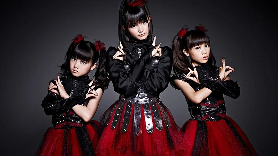 Three Babymetal members in black and red gothic stage outfits posing against a dark background