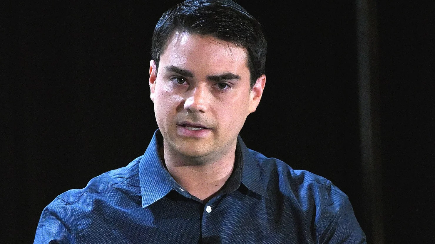 Portrait of Ben Shapiro in a dark blue shirt against a black background