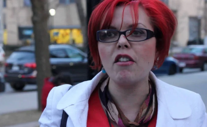 Big Red — Red-haired woman in a white jacket and scarf speaking on a city sidewalk during an interview