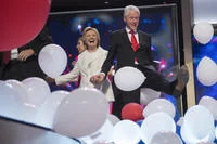 Bill Clinton Loves Balloons — Hillary Clinton in a white outfit and Bill Clinton in a dark suit stand together on a brightly lit DNC stage, grinning as red
