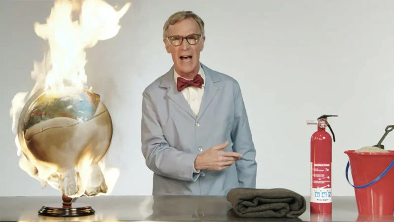 Bill Nye The Planet Is On Fucking Fire Video — Bill Nye wearing his signature blue shirt and red bow tie stands beside a flaming globe and fire extinguisher on