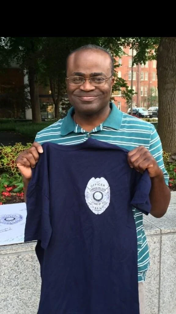 Black Guy On The Phone — Smiling Black man in teal striped polo holding up a navy t-shirt supporting Officer Darren Wilson