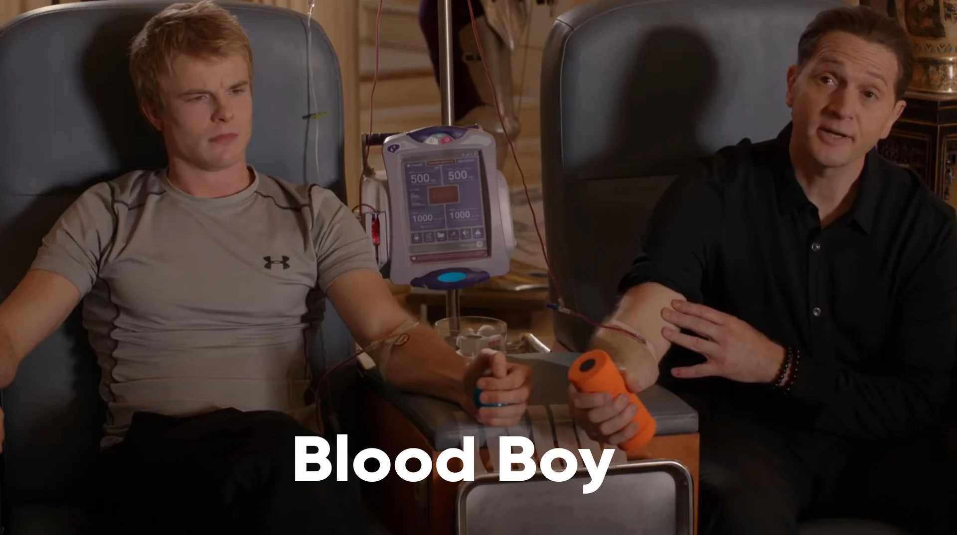 Young man and older man seated in chairs connected by a blood transfusion machine, labeled 'Blood Boy'.