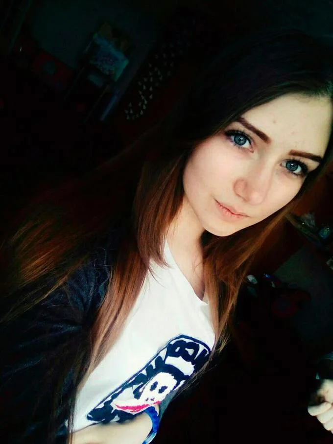 Blue Whale Challenge — A selfie of a young woman with long auburn-dark hair and blue eyes wearing a white graphic t-shirt, photographed in a dimly lit room.