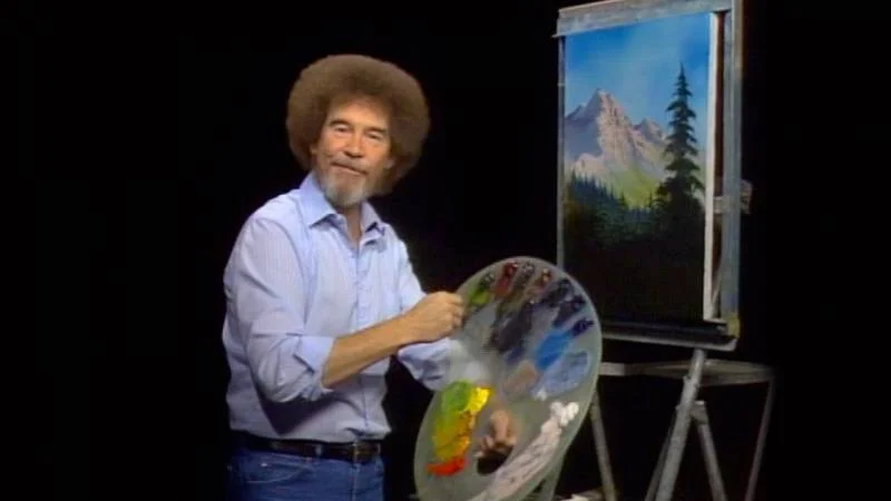 Bob Ross holding a paint palette in front of a mountain landscape painting on an easel