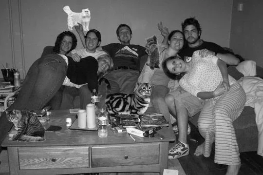 Boozecats — A black and white photograph of people on a couch with several cats digitally inserted throughout the scene, positioned as replacements for drinks.