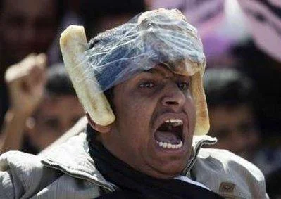 Bread Helmet Man — A tight close-up photograph of a protester wearing two bread loaves strapped to his head with plastic wrap, mouth wide open in a shout, with
