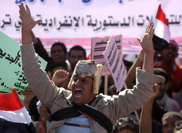 A wide press photograph of Bread Helmet Man shouting with both arms raised, wearing bread loaves on his head, with colorful Arabic protest banners and Yemeni