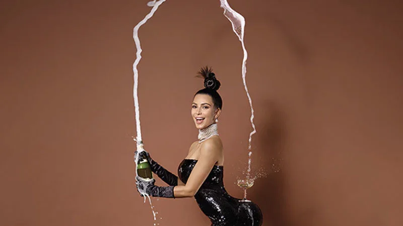 Break The Internet — Kim Kardashian poses in black sequined strapless outfit and long gloves against tan background, balancing champagne coupe on rear with