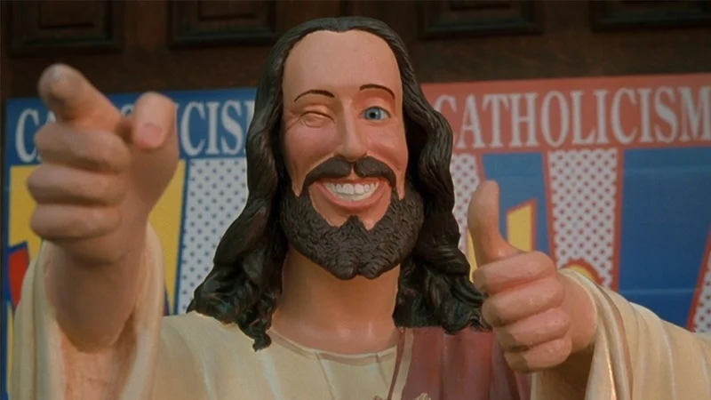 Buddy Christ statue winking and giving a thumbs-up in front of a Catholicism banner