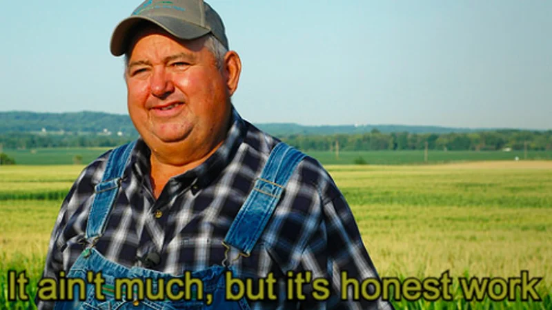 But Its Honest Work — Smiling farmer in denim overalls and cap standing in a field with the caption 'It ain't much, but it's honest work'