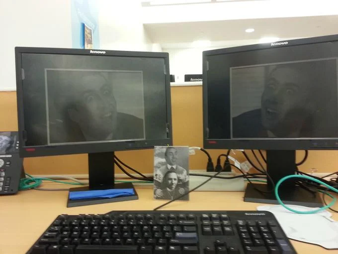 Caging — Two office monitors showing Nicolas Cage screensavers with a framed Cage photo on the desk