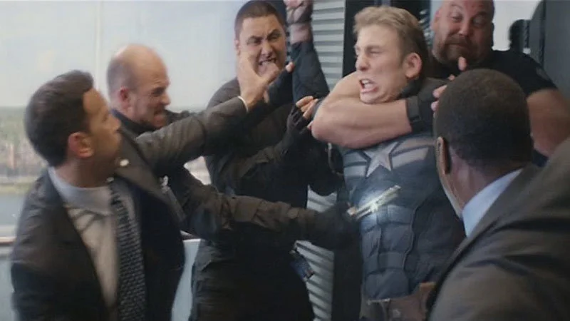 Captain America Elevator Fight — A clean movie still from Captain America: The Winter Soldier showing Steve Rogers grappling with suited men in an elevator.