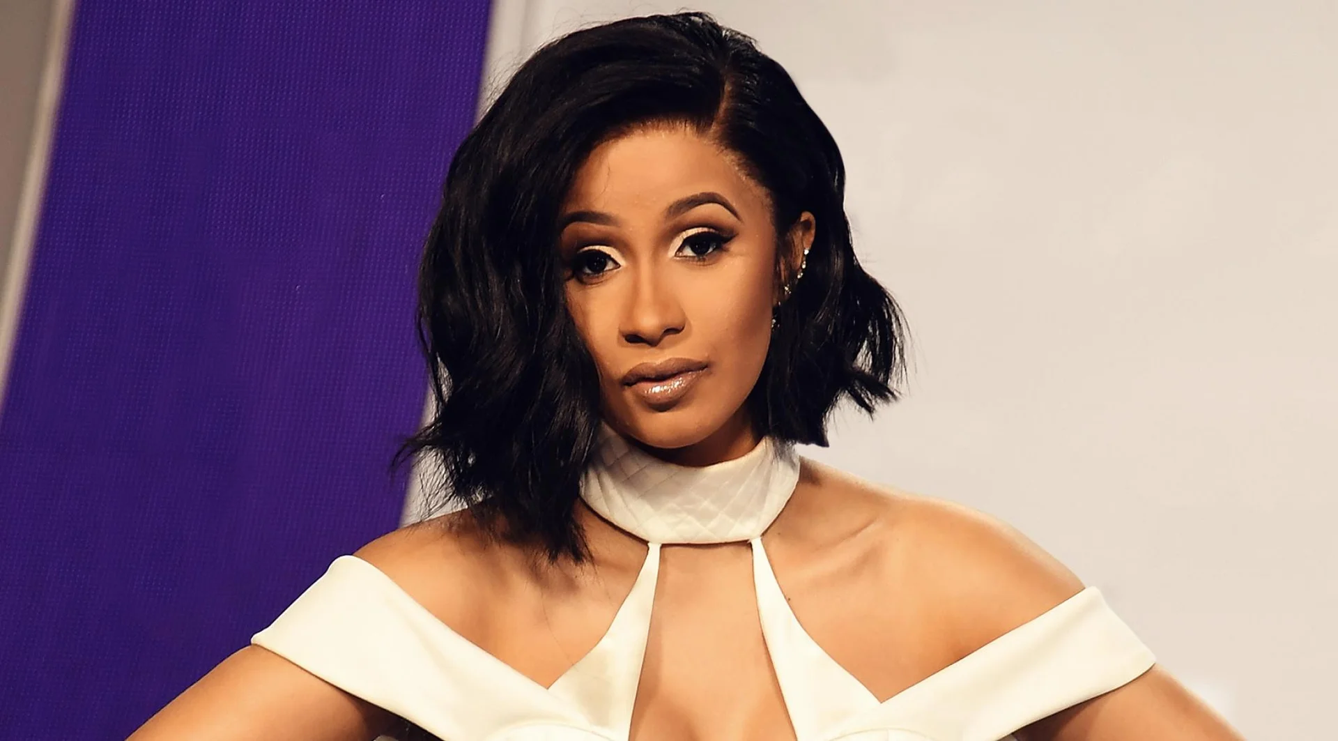 Cardi B posing on the red carpet in a white off-shoulder outfit with a purple backdrop