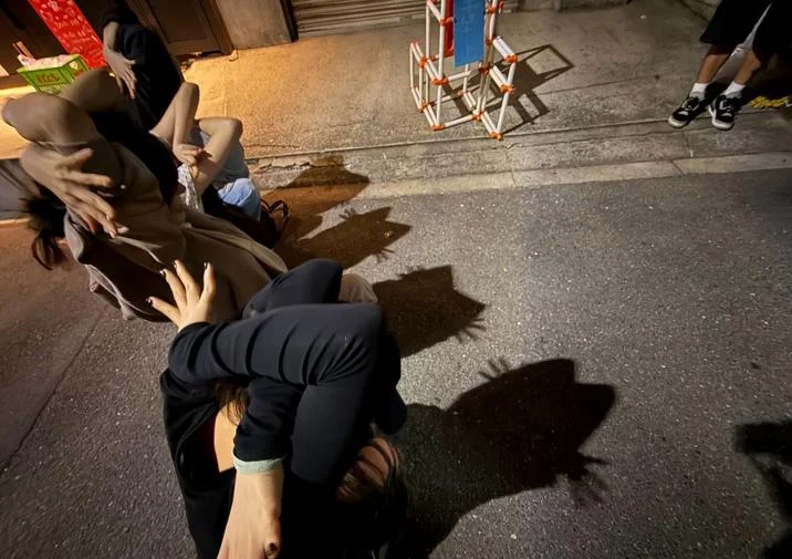 Two people crouch together on a busy nighttime sidewalk performing the Cat Shadow Trend with bystanders nearby.