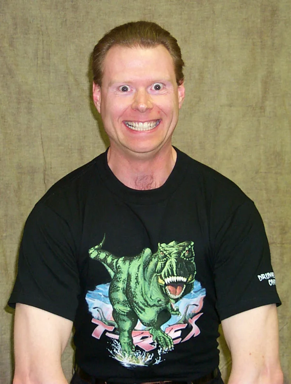 Cockmongler — Smiling man with wide eyes wearing a green T-Rex print black t-shirt