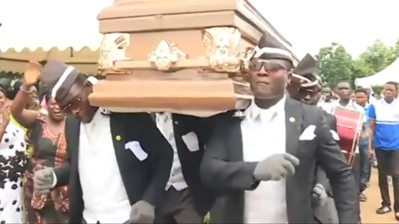Pallbearers in dark formal suits and hats carry a wooden coffin while performing the Coffin Dance in an outdoor funeral setting with spectators.