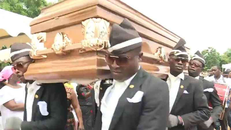 Pallbearers in formal dark suits and hats perform the Coffin Dance while carrying a wooden coffin in an outdoor setting with spectators and trees.