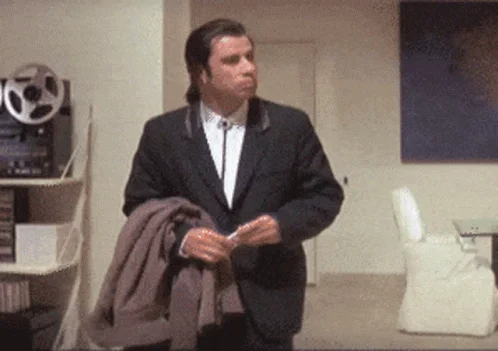 A still frame from the original Confused Travolta GIF showing Travolta in a black suit carrying a jacket and looking around with a puzzled expression.