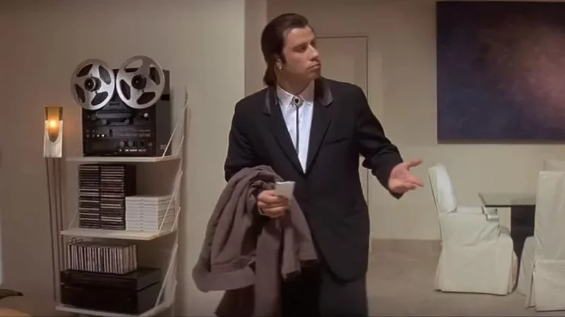 Confused Travolta — John Travolta as Vincent Vega in his iconic confused pose wearing a black suit and expressing bewilderment with open hand gestures in a