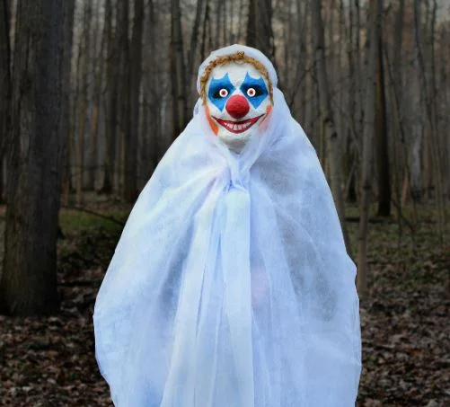 Creepy Clown Sightings — Person in creepy clown mask and white sheet standing in wooded area