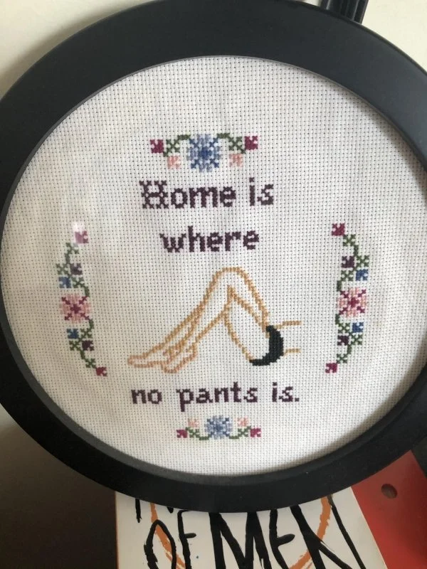 Cross-Stitch Art — Cross-stitch hoop with floral designs and text 'Home is where no pants is'
