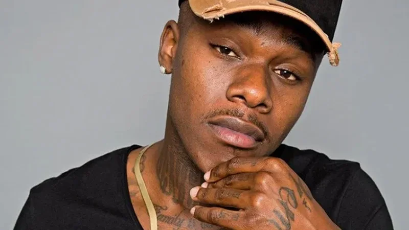 Professional portrait of DaBaby wearing gold/orange baseball cap and black shirt with signature hand-to-chin pose.