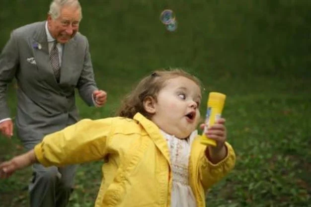 Dancing Prince Charles — King Charles III in gray suit playing and dancing with young child in bright yellow jacket holding bubble wand.