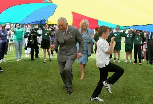 Dancing Prince Charles — King Charles III in gray suit dancing energetically with child in athletic wear at outdoor community event with crowds and umbrellas.